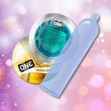 A blue condom beside two circular wrappers labeled "ONE FlavorWaves" and "Banana Split" sits against a pink and purple bokeh backdrop, highlighting ONE Banana Split Flavored Condoms 🍌—vegan, ultra-thin latex condoms by ONE.