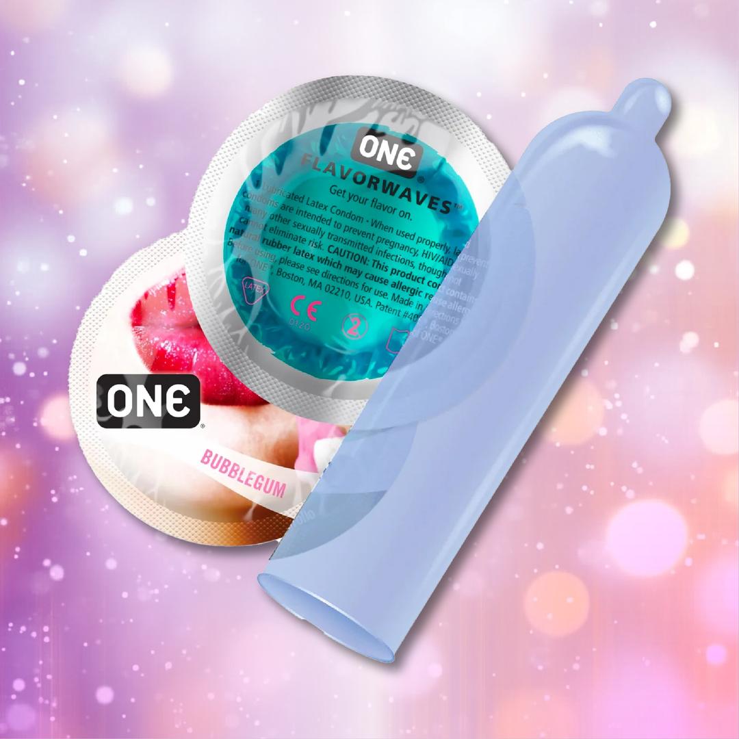 A purple, ultra-thin ONE Bubblegum Flavored Condom stands upright before two circular wrappers labeled "FLAVORWAVES" and "BUBBLEGUM," set against a soft pink and purple bokeh background, highlighting the ONE brand's bubblegum condoms.