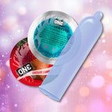 ONE Chocolate Strawberry Flavored Condoms 🍓 by ONE are shown: a red “Chocolate Strawberry” wrapper, a teal “Flavorwaves” wrapper, and an unrolled translucent condom, all set against a pink and purple bokeh background. Vegan-friendly.