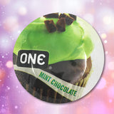 A chocolate cupcake with green mint frosting and chocolate shavings sits on a circular label reading "ONE" and "Mint Chocolate," inspired by the flavor of ONE "Mint Chocolate" Flavored Condoms, against a pink and purple bokeh background.
