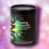 A black cylindrical container labeled "ONE Glowing Pleasure" glow-in-the-dark condoms by ONE, featuring colorful accents, is shown against a pink and purple bokeh background. The package notes 36 natural rubber latex condoms with softer latex.