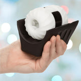 A hand holds the CalExotics Optimum Power Power Thruster Male Masturbator, a black rechargeable device featuring a white textured stroker sleeve with a hollow center. The softly blurred background is filled with pastel colors and lights.