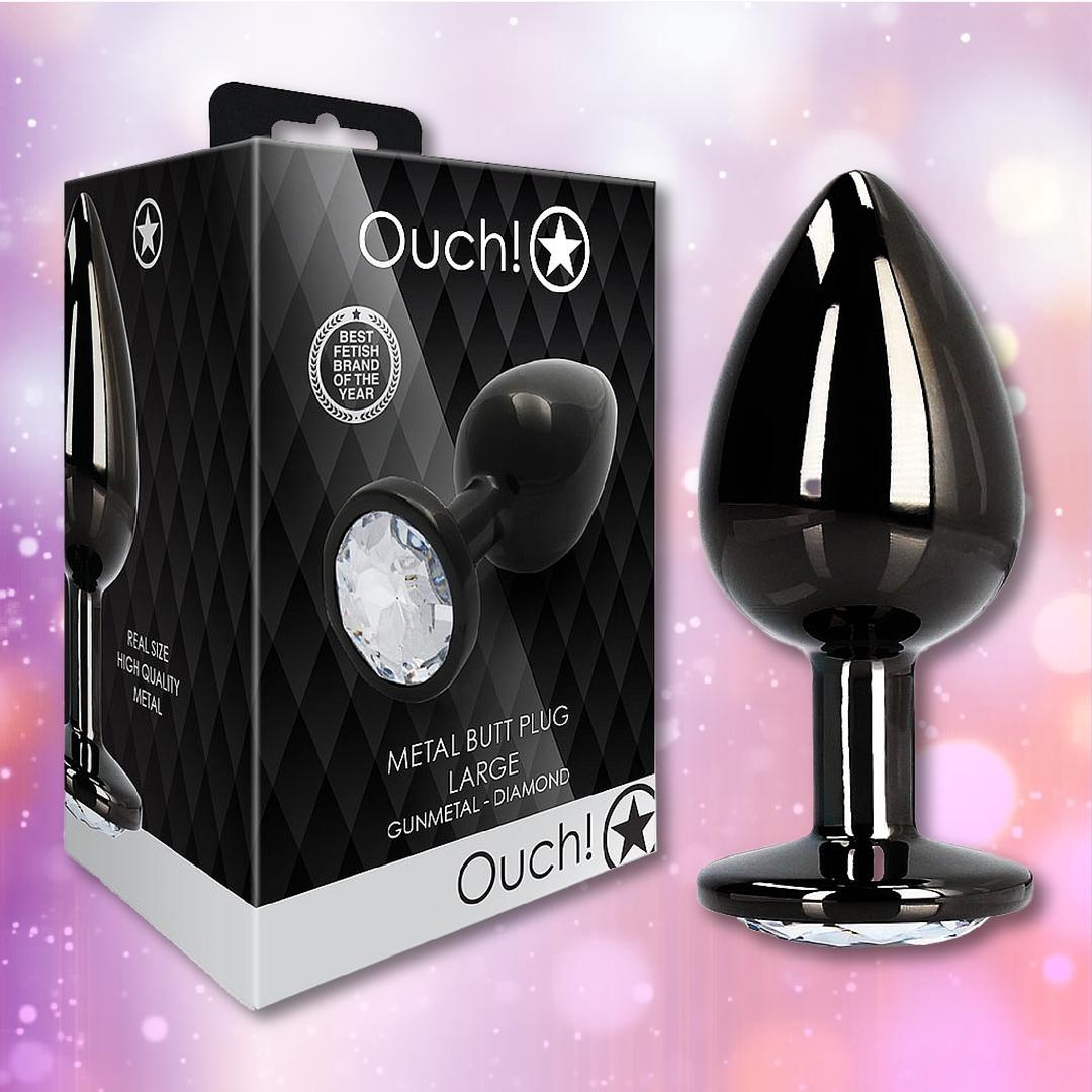 A Ouch! Large Metal Butt Plug – Gunmetal with Diamond Base by Shots stands beside its black, diamond-patterned box. The shimmering pastel background highlights the plug’s sparkling gem base.