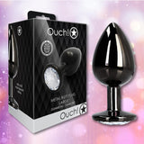 A Ouch! Large Metal Butt Plug – Gunmetal with Diamond Base by Shots stands beside its black, diamond-patterned box. The shimmering pastel background highlights the plug’s sparkling gem base.