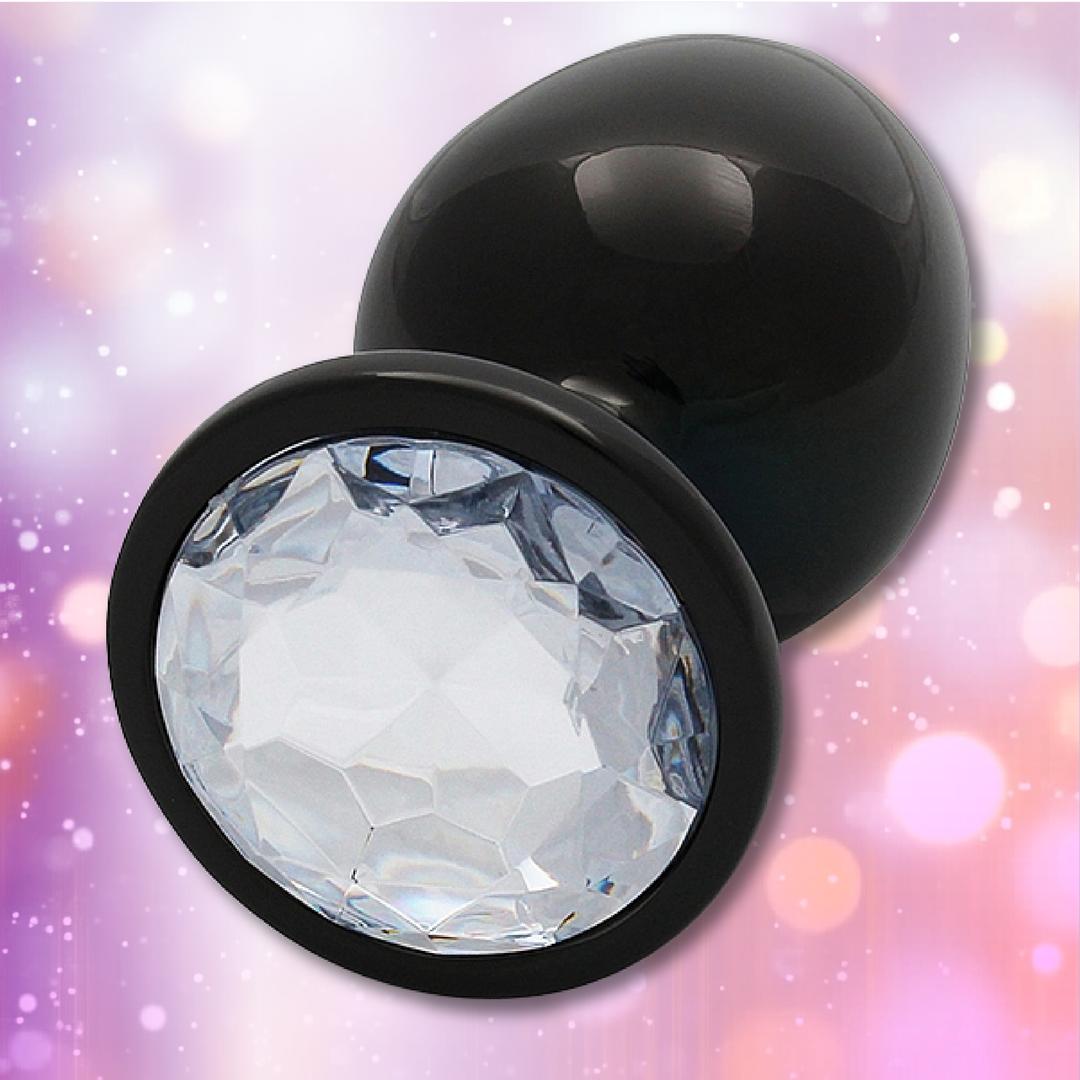 The Ouch! Large Metal Butt Plug by Shots features a sleek, gunmetal finish with a diamond base, displayed against a soft pink and purple bokeh background.