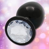 The Ouch! Large Metal Butt Plug by Shots features a sleek, gunmetal finish with a diamond base, displayed against a soft pink and purple bokeh background.