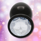 The Ouch! Large Metal Butt Plug by Shots features a sleek gunmetal finish and a clear diamond base, set against a softly glowing pink and purple bokeh background.