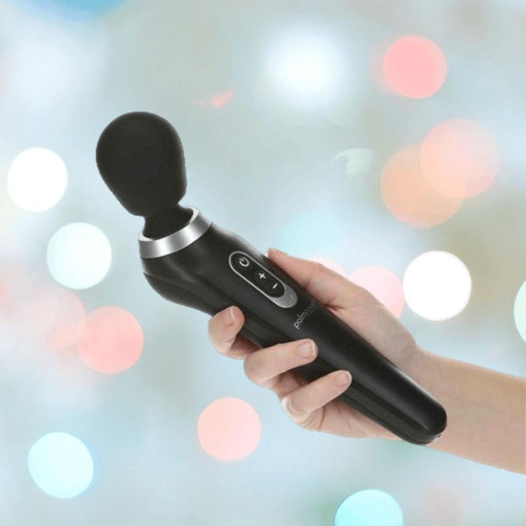 A hand holds the BMS PalmPower Extreme Wand Massager in black, a powerful silicone vibrator with control buttons, against a soft, colorful bokeh background.