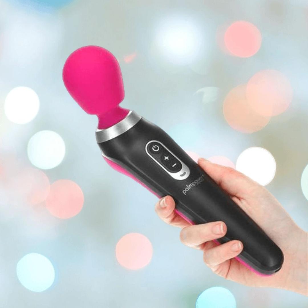 A hand holds the BMS PalmPower Extreme Wand Massager - Pink, featuring control buttons and a smooth silicone finish, against a blurred background with pastel lights.