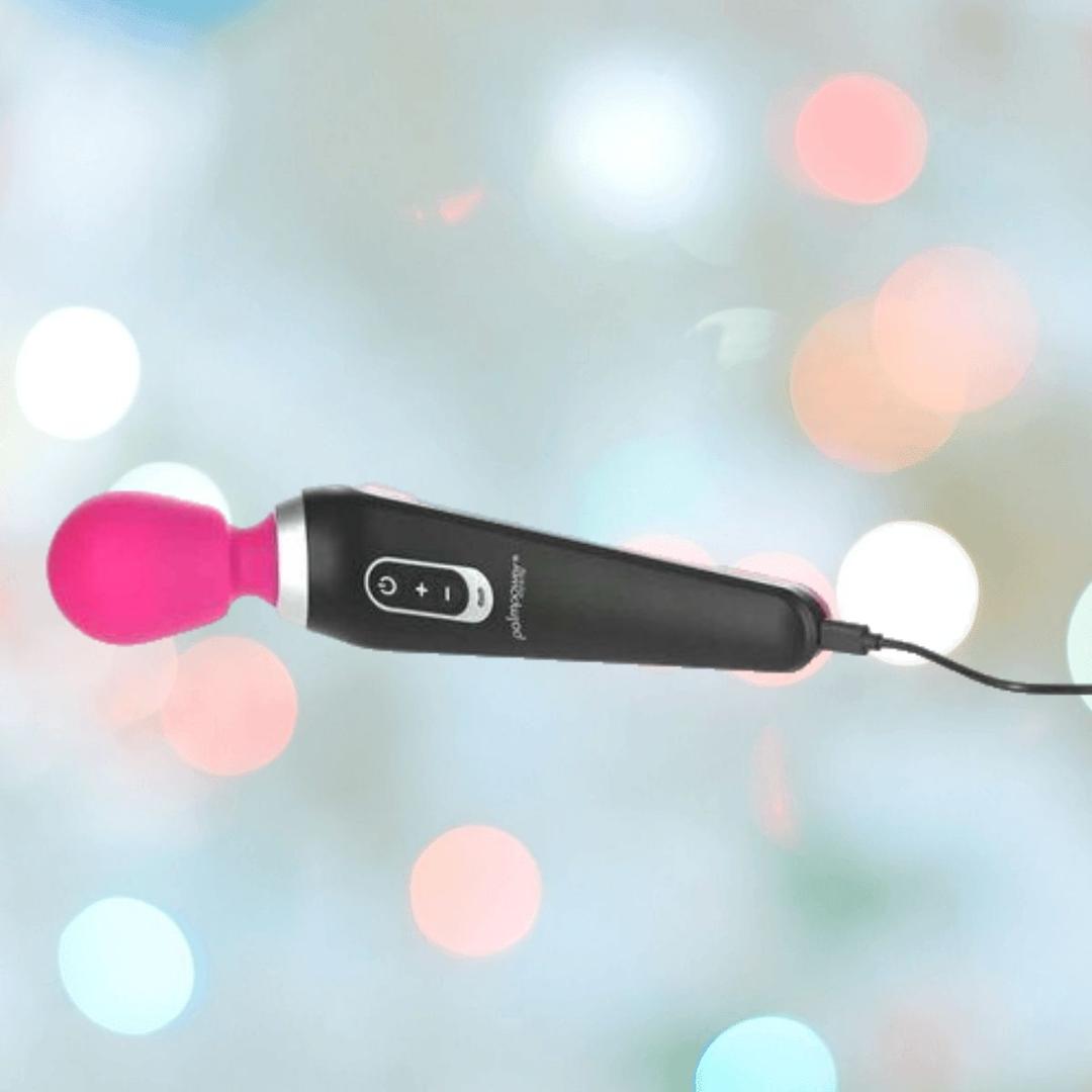 The BMS PalmPower Extreme Wand Massager in pink features a black rechargeable handle, a vibrant pink silicone head, and control buttons. It’s shown plugged in against a softly blurred backdrop of colorful lights.