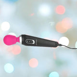 The BMS PalmPower Extreme Wand Massager in pink features a black rechargeable handle, a vibrant pink silicone head, and control buttons. It’s shown plugged in against a softly blurred backdrop of colorful lights.