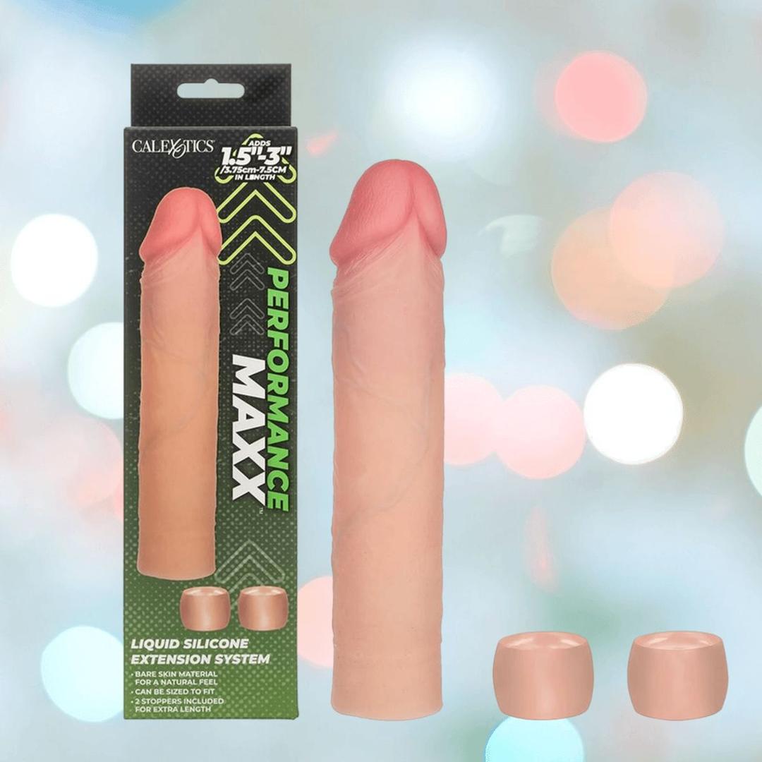 The CalExotics Performance Maxx Liquid Silicone Extension System - Ivory, shown with two extra silicone ring caps, is displayed beside its green and black box amid softly blurred colorful lights.
