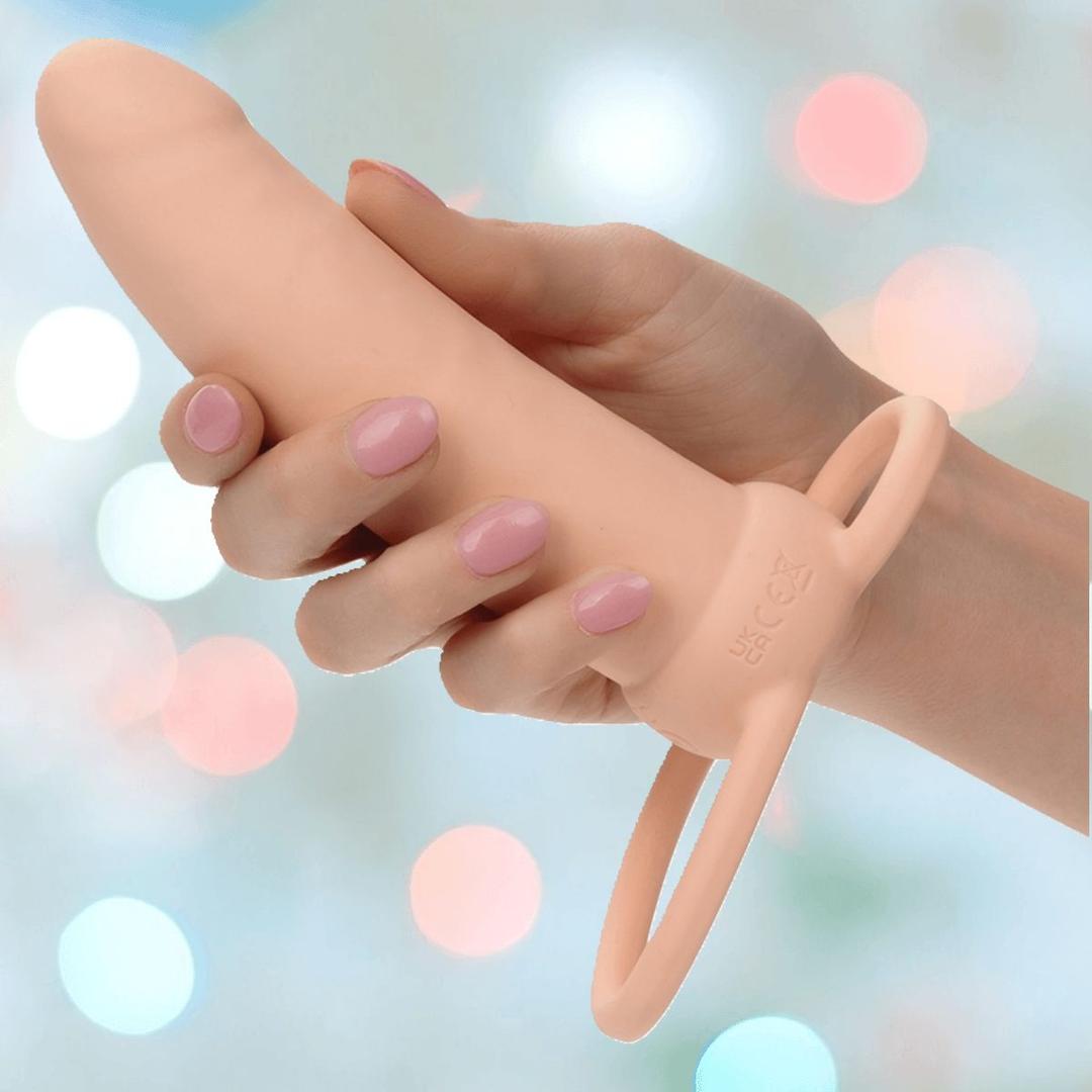 A manicured hand holds the CalExotics Performance Maxx Rechargeable Thick Dual Penetrator in ivory, featuring two looped handles, against a softly blurred pastel background.