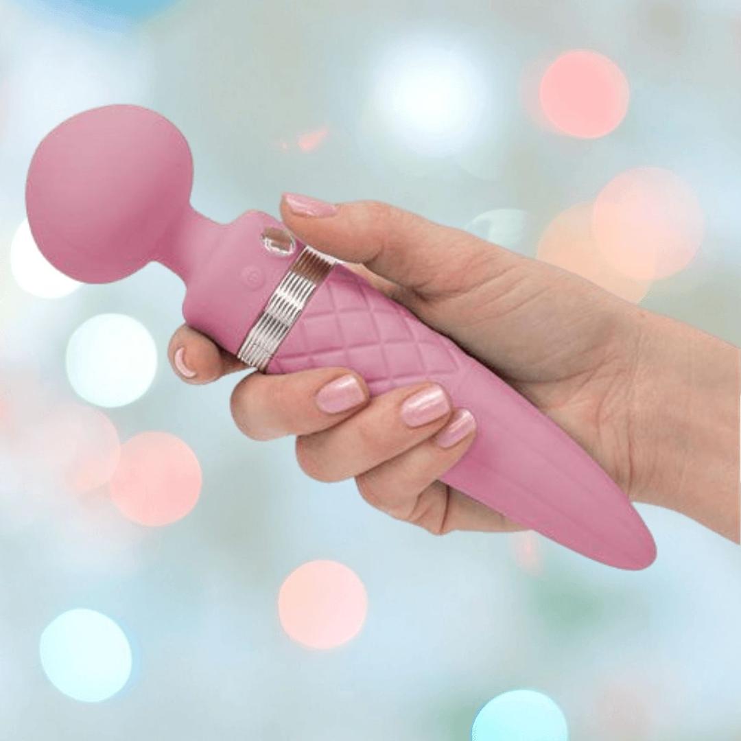 A hand holds the BMS Pillow Talk Sultry Wand Massager in pink, featuring a quilted handle and rounded head for G-spot stimulation, set against a softly blurred pastel background.