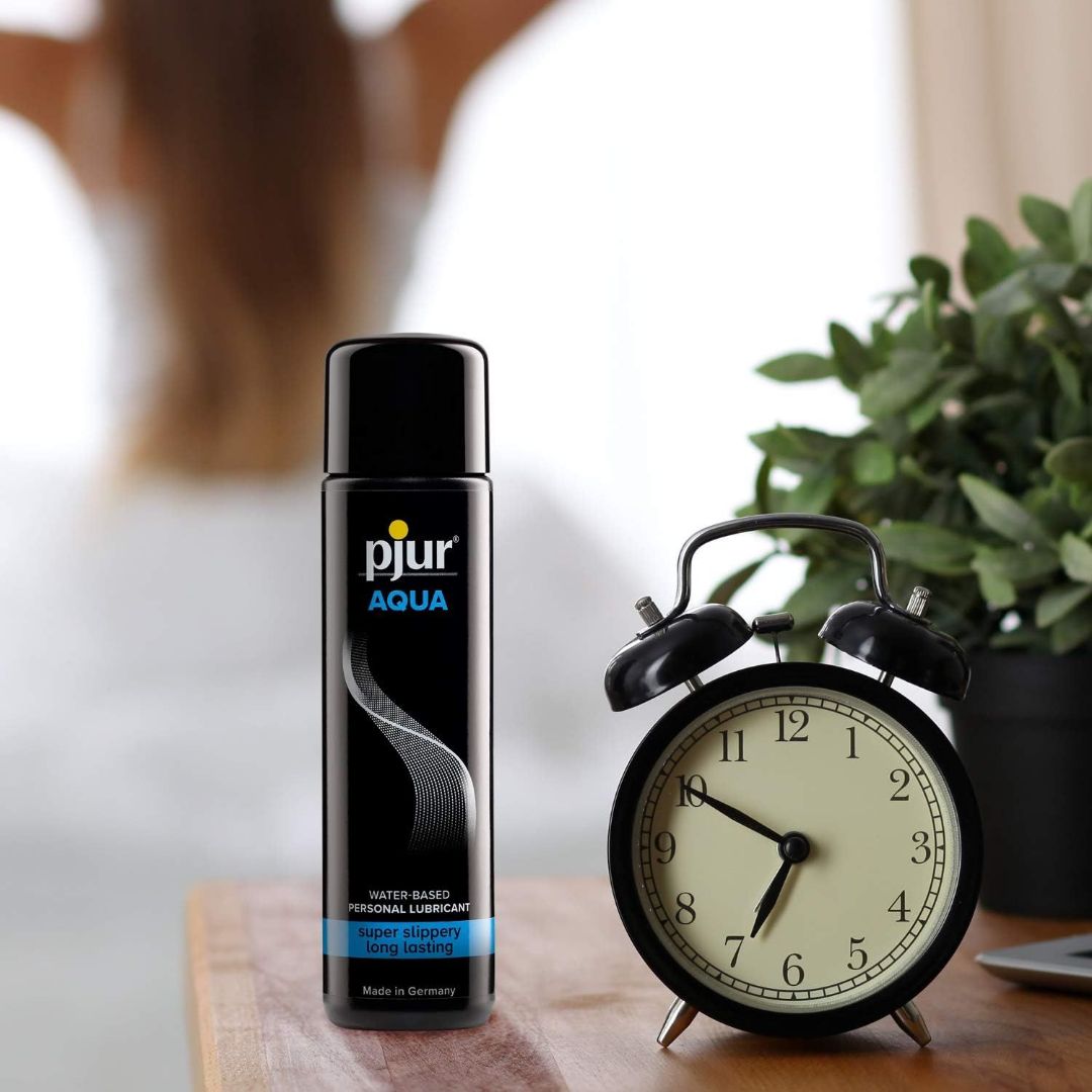 A 100ml (3.4oz) bottle of Pjur AQUA Water-Based Lubricant is placed next to a black retro alarm clock and a small potted plant. In the blurred background, there is a person with long hair sitting.