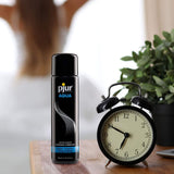 A 100ml (3.4oz) bottle of Pjur AQUA Water-Based Lubricant is placed next to a black retro alarm clock and a small potted plant. In the blurred background, there is a person with long hair sitting.