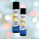 Two bottles of Pjur Analyse Me! Comfort Anal Glide Lubricant (Water-Based), featuring blue and yellow accents, are positioned against a backdrop of blurred colorful lights. The larger bottle holds 250 ml/8.5 fl oz, while the smaller one contains 100 ml/3.4 fl oz.