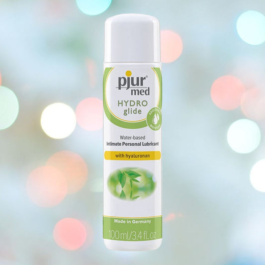 The image features a 100ml (3.4 oz) bottle of Pjur Med Hydro Glide, a water-based lube by Pjur that is hypoallergenic and enriched with moisturizing Hyaluronan. The bottle is white with a green and yellow label, placed against a softly focused multicolored bokeh background. 1080