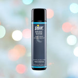 Bottle of Pjur "Basic" Water-Based Lubricant | 100ml (3.4oz), an affordable personal lubricant, set against a blurred colorful background. The blue and black bottle's label emphasizes its compatibility with latex condoms and gentle formulation for sensitive skin, proudly made in Germany.