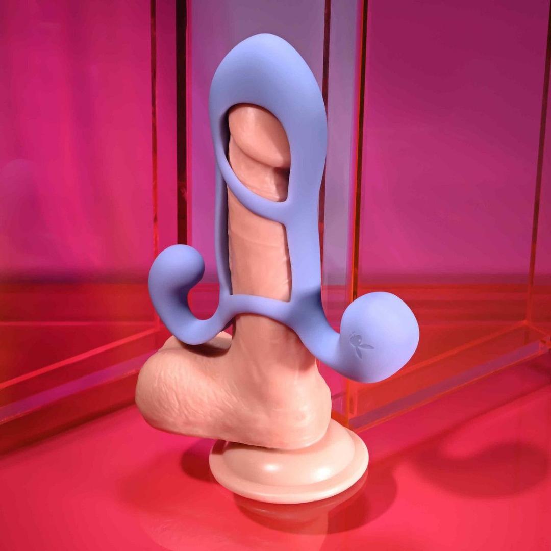 The Playboy Fantasy Cage by Evolved Novelties is a realistic dildo with a blue silicone attachment featuring ring and arm extensions, shown upright on a pink surface against a vibrant pink and orange background.