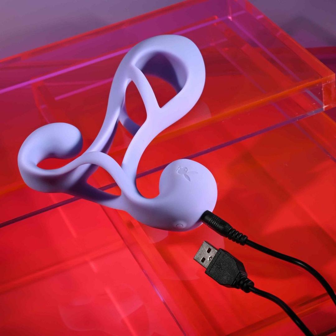 The Playboy Fantasy Cage by Evolved Novelties, a purple silicone adult toy with an abstract curved design, is connected to a USB charging cable and rests on a transparent red and purple acrylic surface.