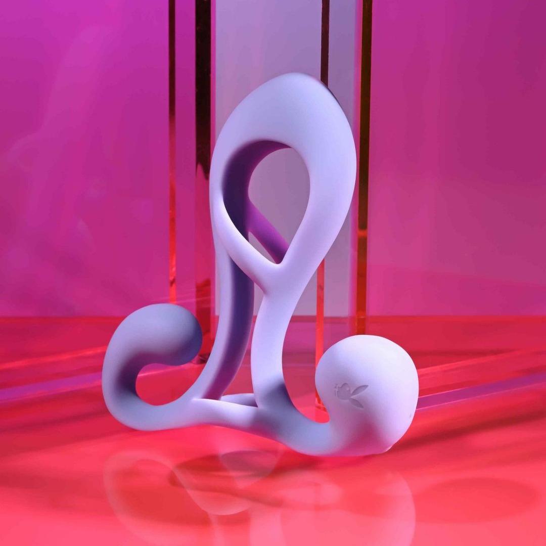 The Playboy Fantasy Cage by Evolved Novelties is a modern, abstract sculptural object in lavender with smooth curves and circular ends, displayed against a vibrant pink and red reflective background.