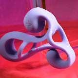 The Playboy Fantasy Cage by Evolved Novelties is a glossy, abstract lavender sculpture with swirling, twisted loops and a fluid form, displayed against a vibrant pink and red gradient background.