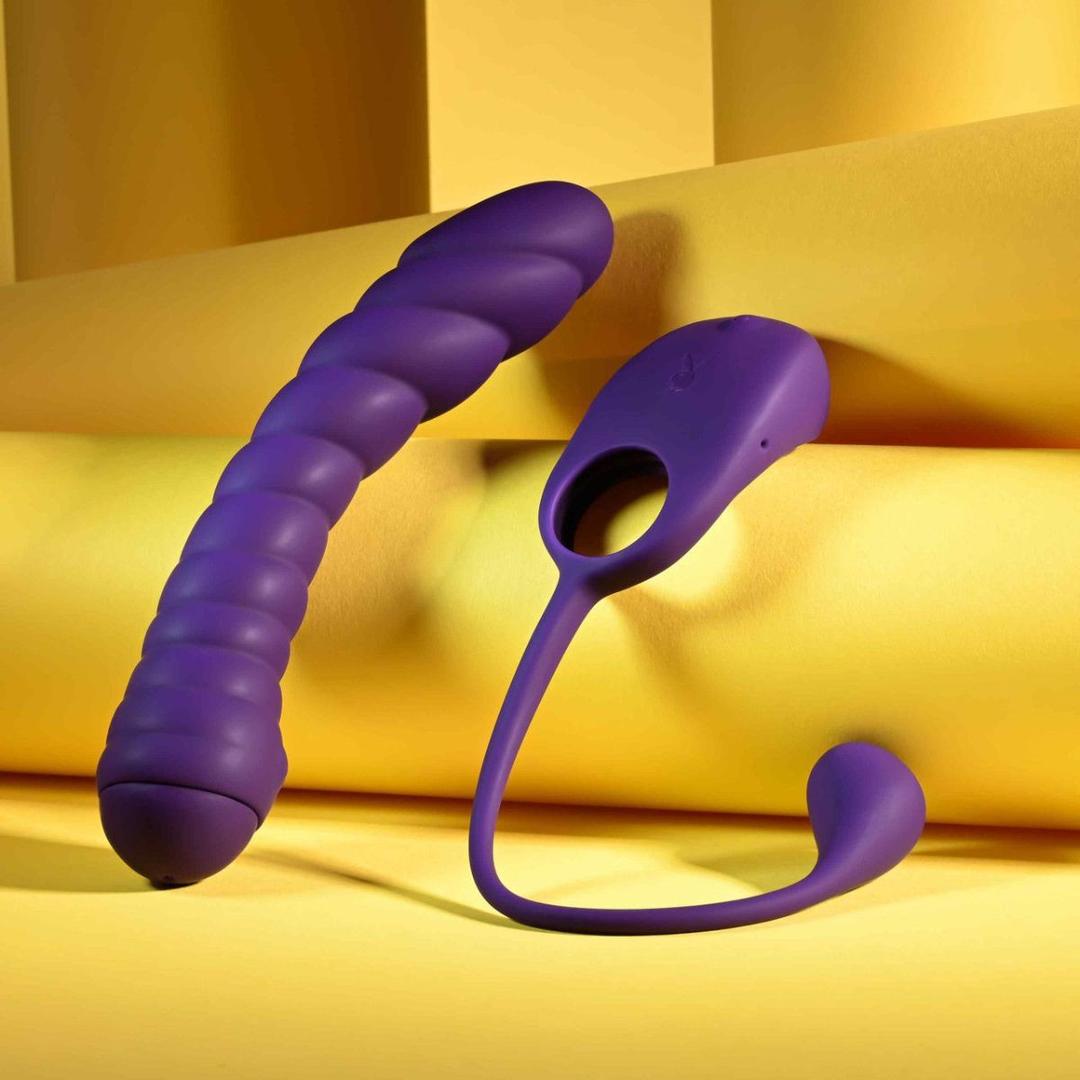 Two Playboy Twisted Playground toys by Evolved Novelties stand upright on yellow fabric. One is ribbed and curved, the other features a ring handle and long flexible arm; both have a smooth, modern look.