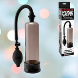 The Pipedream "Pump Worx Beginners Power Penis Pump - Black," designed to enhance erections, includes a transparent cylinder and a black rubber hose, displayed next to its packaging. The box highlights the product name against a softly lit, blurred background.