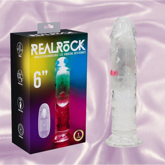 The RealRock Wireless Rechargeable LED Vibrating 6" by Shots America, featuring a clear body, remote control, and sturdy suction cup, is displayed beside its vibrant packaging on a shiny light fabric background. 1080