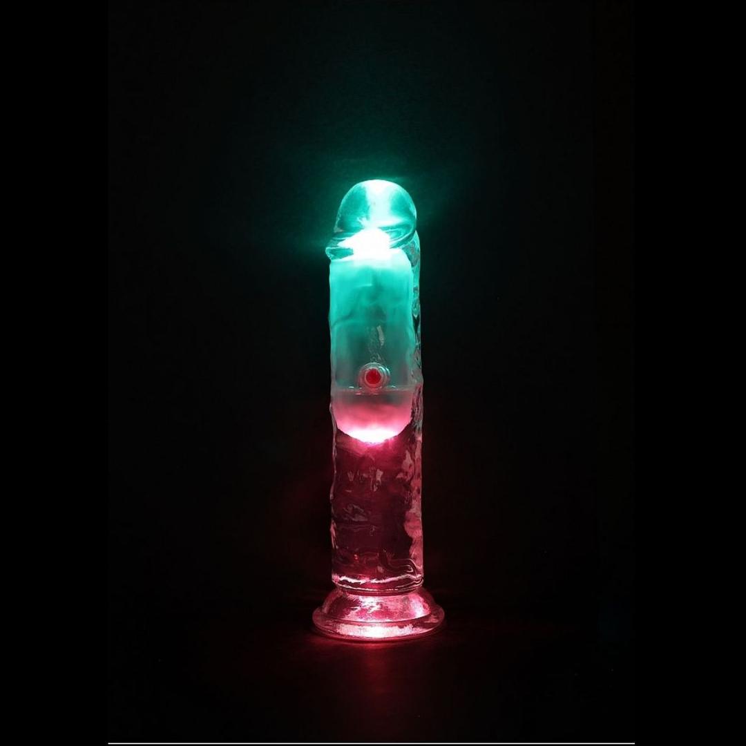 The RealRock Wireless Rechargeable LED Vibrating 6" by Shots America, a transparent cylindrical dildo, glows with pink and teal lights from below, creating a vibrant effect in the dark.