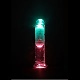 The RealRock Wireless Rechargeable LED Vibrating 6" by Shots America, a transparent cylindrical dildo, glows with pink and teal lights from below, creating a vibrant effect in the dark.