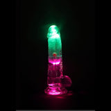 The RealRock Wireless Rechargeable LED Vibrating With Balls 6" by Shots America is a transparent dildo illuminated from within by green and pink lights, shown against a black background.