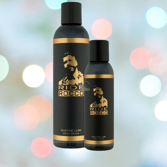 Two black bottles of Sliquid's "Ride Bodyworx Ride Rocco Silicone-Based Lube" with gold accents sit against a blurred background of multicolored lights. One bottle is larger and the other is smaller, both featuring a silhouette of a bearded man and the text "RIDE ROCCO". 1080