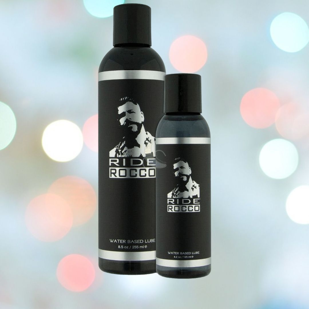 Two bottles of "Ride Bodyworx 'Rocco' Water-Based Lube" by Sliquid are displayed in front of a blurred background with bokeh lights. The larger bottle, holding 250 ml (8.5 oz), and the smaller bottle, containing 125 ml (4 oz), both feature a silhouette image of a man and white text on their black bottles, highlighting their hypoallergenic formula with carrageenan extracts.