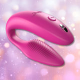 The We-Vibe Sync 2 Couples Vibrator with App in Rose by We-Vibe, featuring a curved design and textured surface on one end, is displayed against a soft, pastel bokeh background.