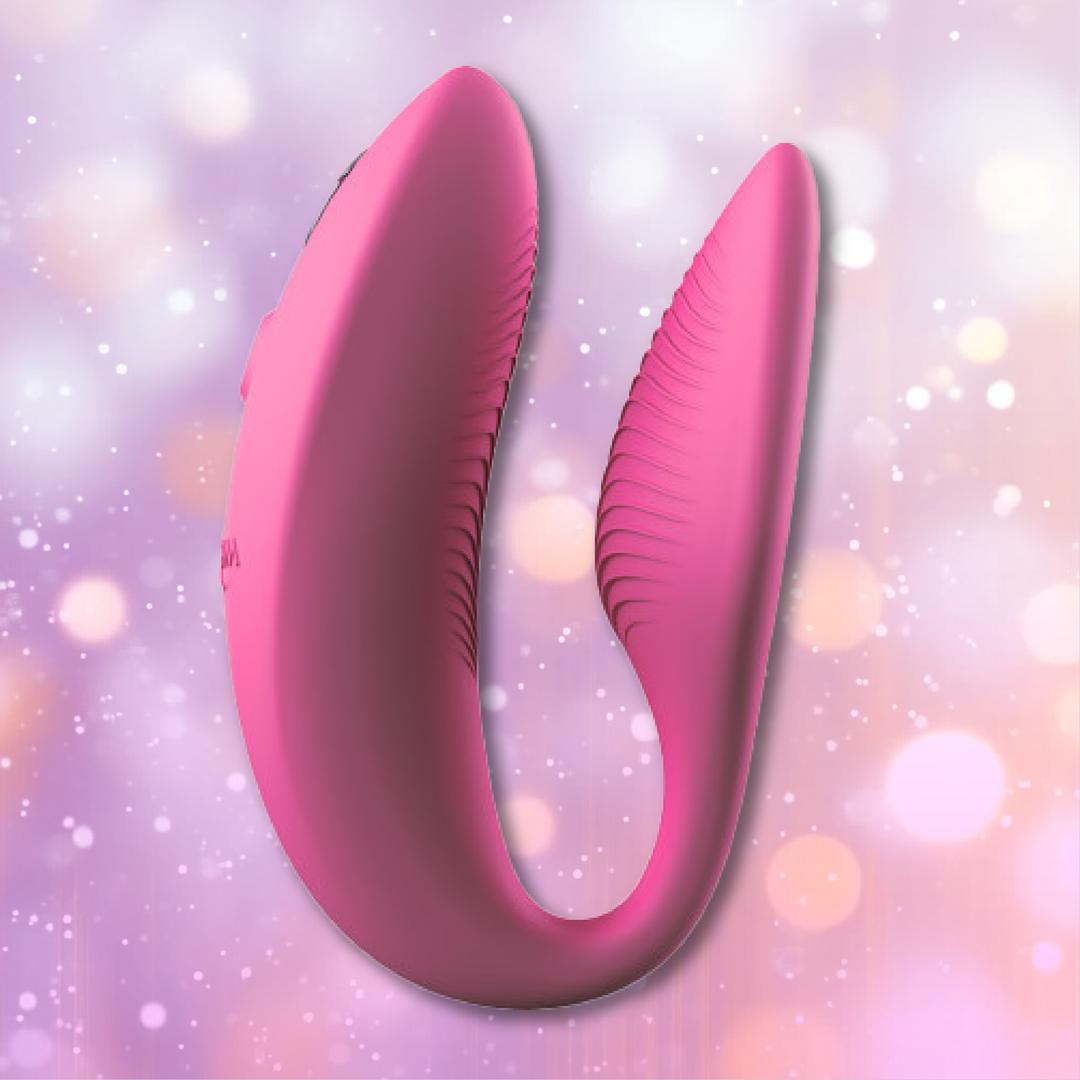 The We-Vibe Sync 2 Couples Vibrator with App in Rose by We-Vibe, a pink U-shaped silicone device with textured sides, is displayed against a pastel bokeh background.