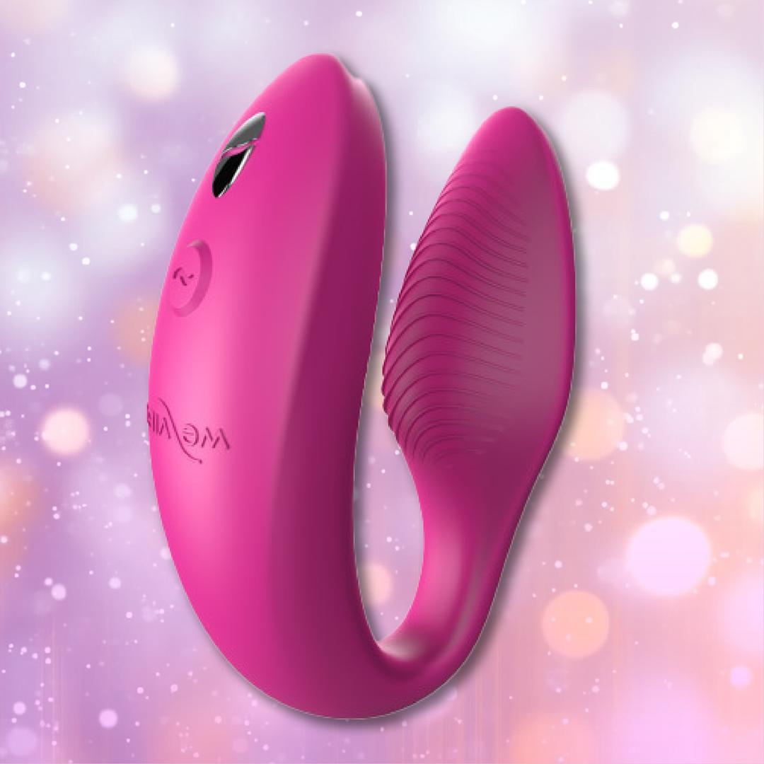 A blush pink, U-shaped couples vibrator with a textured inner surface—the We-Vibe Sync 2 Couples Vibrator with App in Rose by We-Vibe—rests on a softly blurred pastel background highlighted by gentle bokeh, designed for partner play and remote control pleasure.
