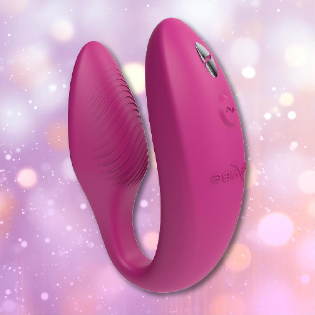 The We-Vibe Sync 2 Couples Vibrator with App - Rose by We-Vibe is a pink, U-shaped silicone couples vibrator featuring buttons on one end, showcased against a soft, pastel bokeh background.