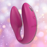 The We-Vibe Sync 2 Couples Vibrator with App - Rose by We-Vibe is a pink, U-shaped silicone couples vibrator featuring buttons on one end, showcased against a soft, pastel bokeh background.