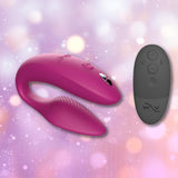 A rose-colored We-Vibe Sync 2 Couples Vibrator with App, featuring a curved and textured design, is displayed alongside a sleek black wireless remote control. Softly blurred purple and peach lights in the background set off the elegance of this We-Vibe creation.