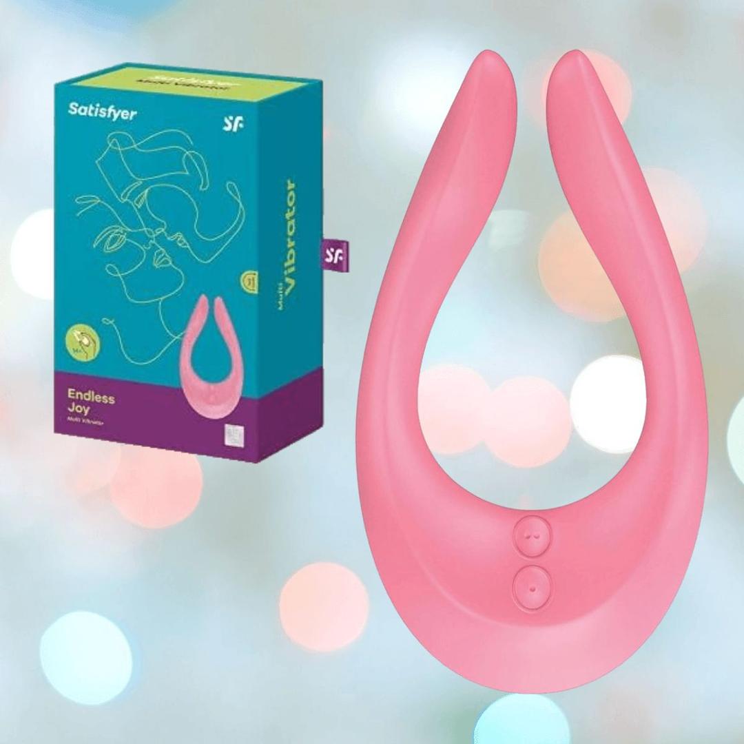 The Satisfyer Endless Joy Vibrator for Couples - Pink by Satisfyer is shown next to its teal and pink packaging, set against a soft, bokeh light background.