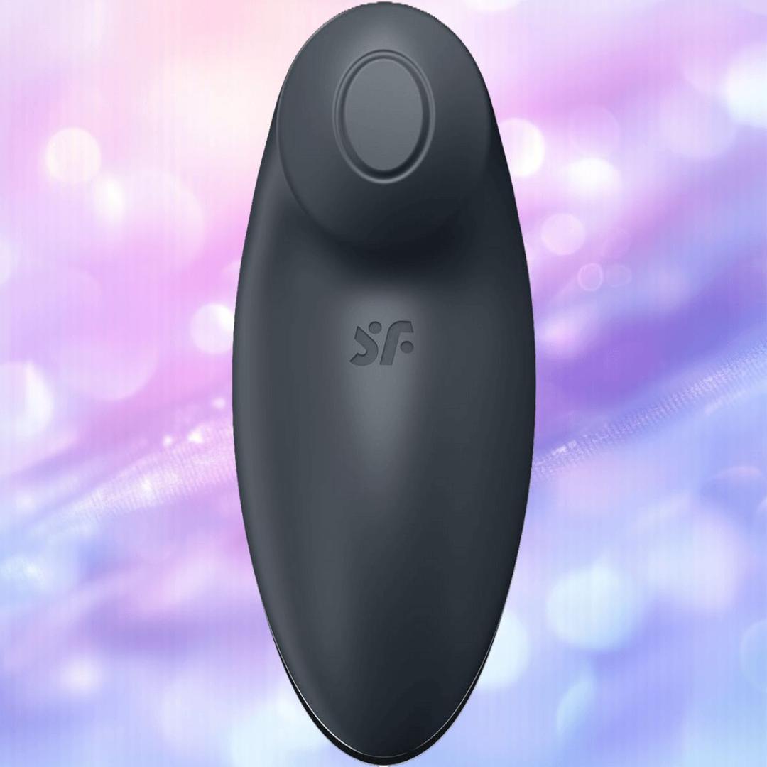 The Satisfyer Tap and Climax 2 Lay-On Vibrator by Satisfyer is a black, oval massager with a round top button and "SF" logo, shown on a softly blurred pastel background in pink, purple, and blue tones.