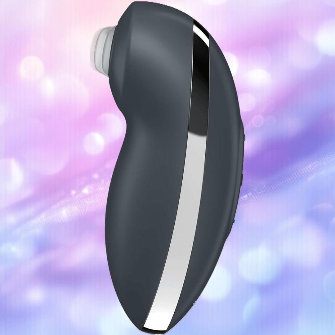 The Satisfyer Tap and Climax 2 Lay-On Vibrator by Satisfyer is a black and silver curved handheld device with a white circular opening at one end, set against a softly blurred pastel purple and blue background.