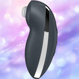 The Satisfyer Tap and Climax 2 Lay-On Vibrator by Satisfyer is a black and silver curved handheld device with a white circular opening at one end, set against a softly blurred pastel purple and blue background.