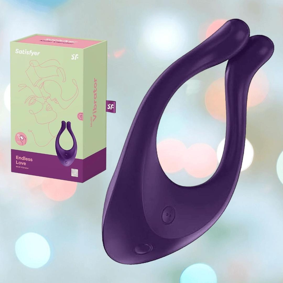 A Satisfyer 'Endless Love' Couples Multi-Use Vibrator in purple is shown before its pastel green and pink packaging, set against a softly blurred background with glowing lights.