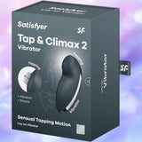 A Satisfyer Tap and Climax 2 Lay-On Vibrator box from Satisfyer, displaying images and details of the toy. The packaging emphasizes its “Sensual Tapping Motion” and a “15 Year Warranty” on a pastel background.