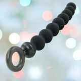 The sleek, black Sportsheets Sex & Mischief Silicone Anal Beads, made from body-safe silicone with a ring handle, invite backdoor exploration against a blurred background lit by soft, colorful bokeh lights.