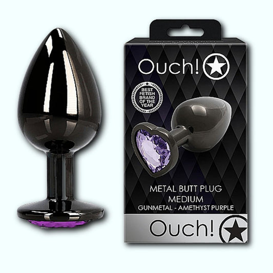 Image of the Shots Ouch! Heart Gem Butt Plug Medium and its packaging. The product features a gunmetal finish with an amethyst purple heart-shaped jewel encrusted base, ideal for temperature play. The packaging showcases the brand "Shots" with the logo "Ouch!" against a diamond-patterned background with a star design. The text reads "Metal Butt Plug Medium. 1080