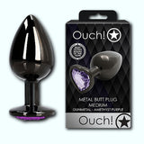 Image of the Shots Ouch! Heart Gem Butt Plug Medium and its packaging. The product features a gunmetal finish with an amethyst purple heart-shaped jewel encrusted base, ideal for temperature play. The packaging showcases the brand "Shots" with the logo "Ouch!" against a diamond-patterned background with a star design. The text reads "Metal Butt Plug Medium.